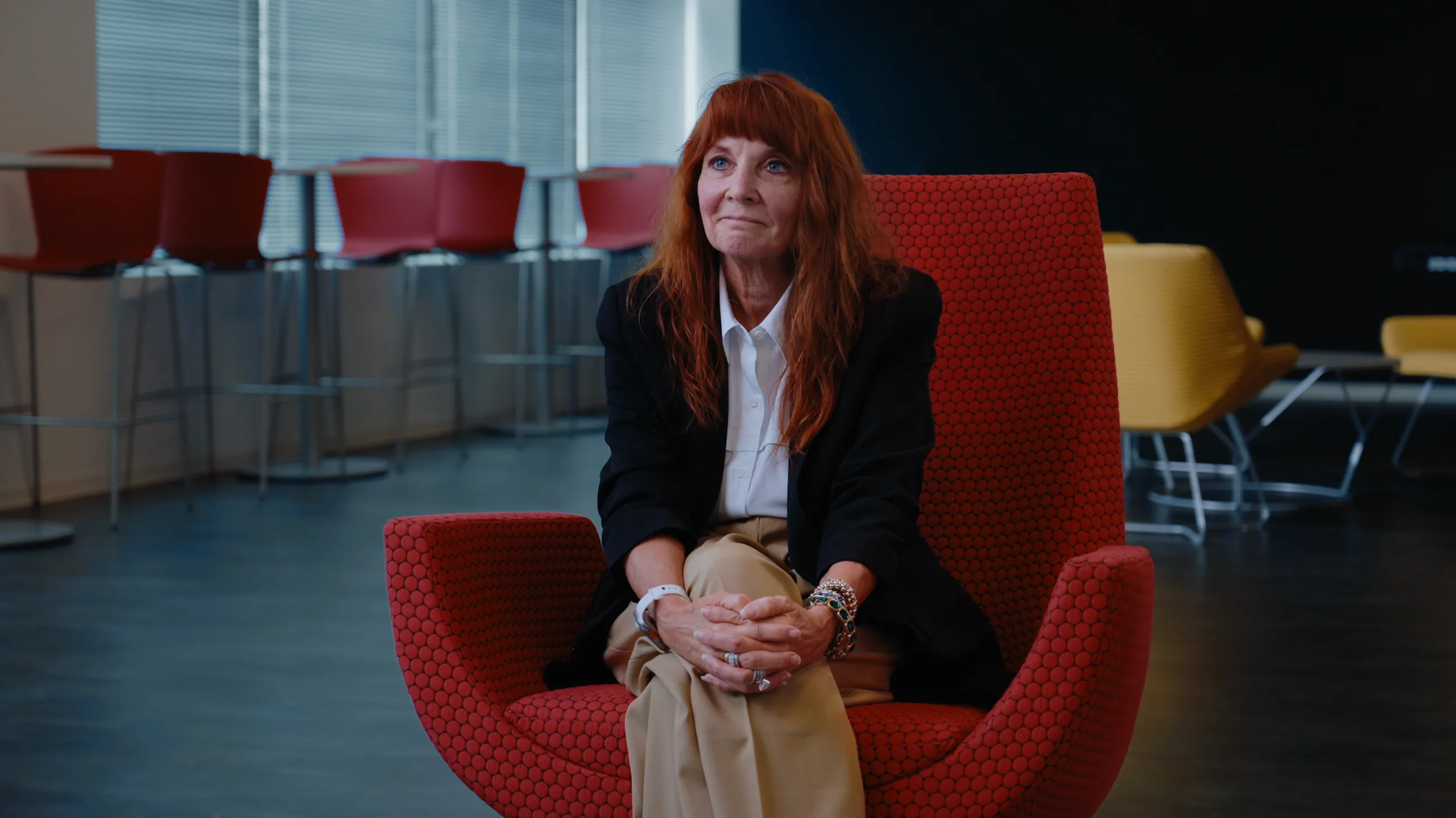 A woman with long auburn hair listens and smiles from a red chair, bar stools and yellow lounge chairs behind her