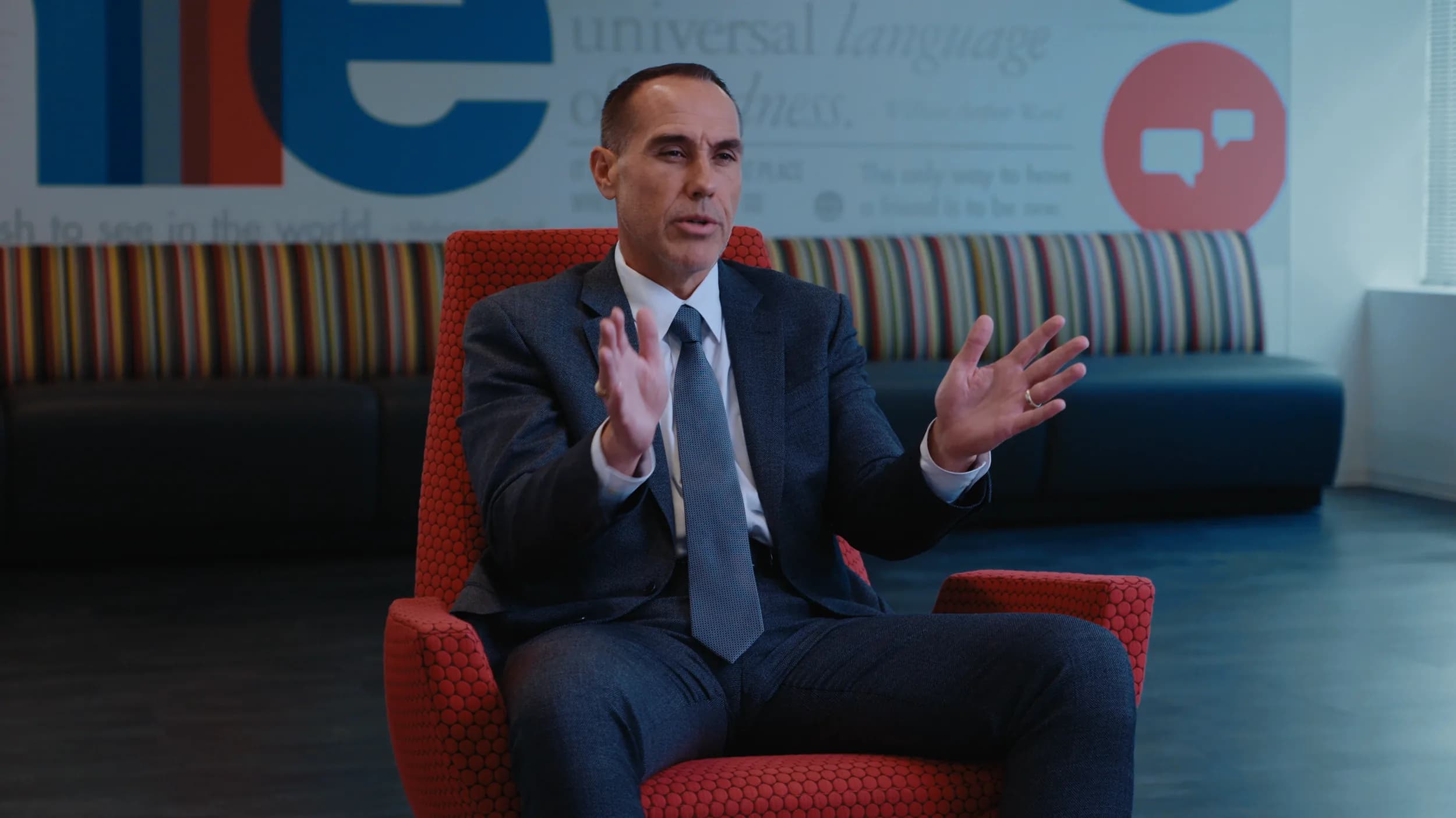 A man in a grey suit speaks with both hands raised from a red chair, a striped bench and mural wall behind him