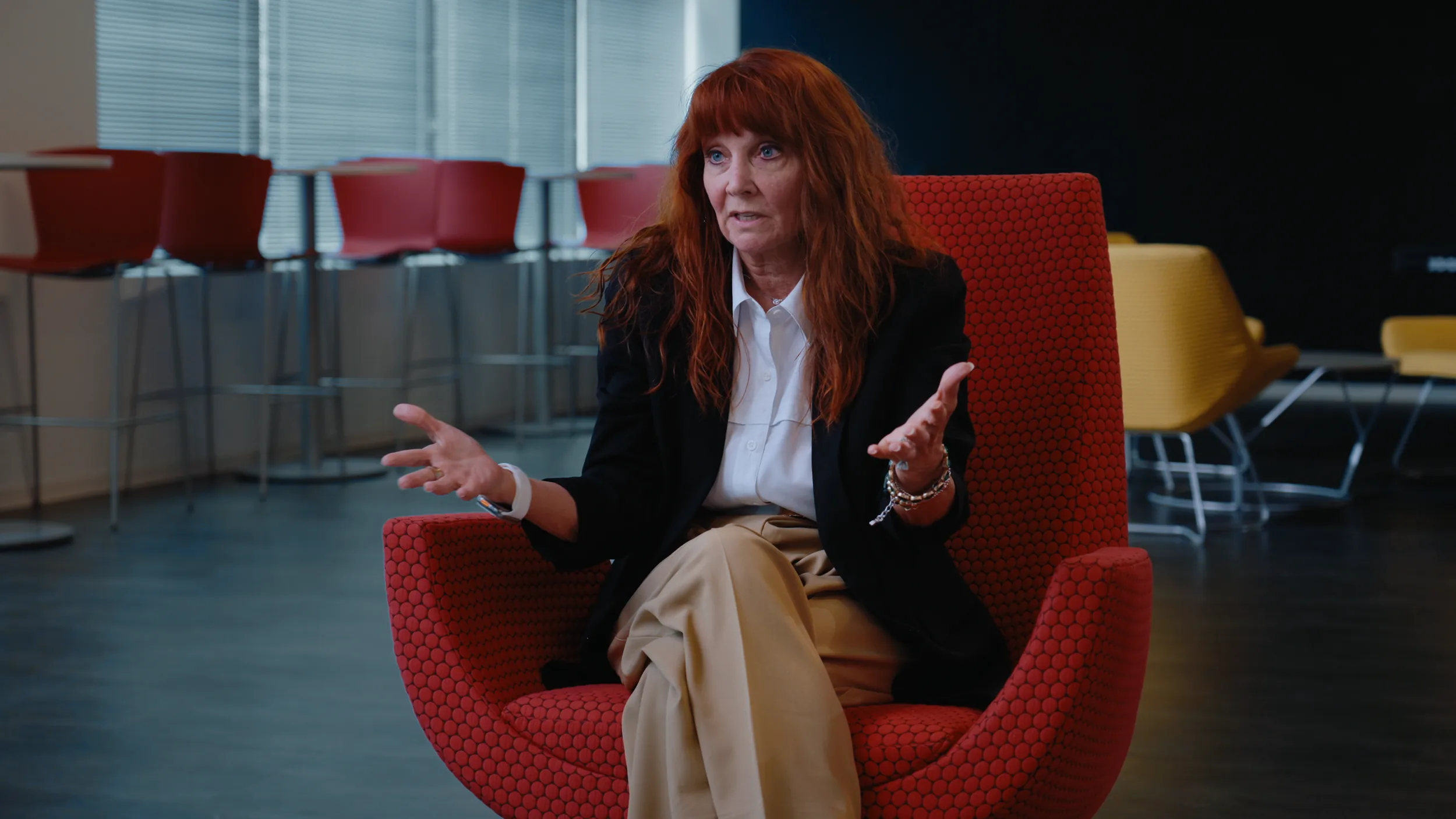 A woman with long auburn hair in a black blazer speaks with open hands from a red chair, bar stools and windows behind her