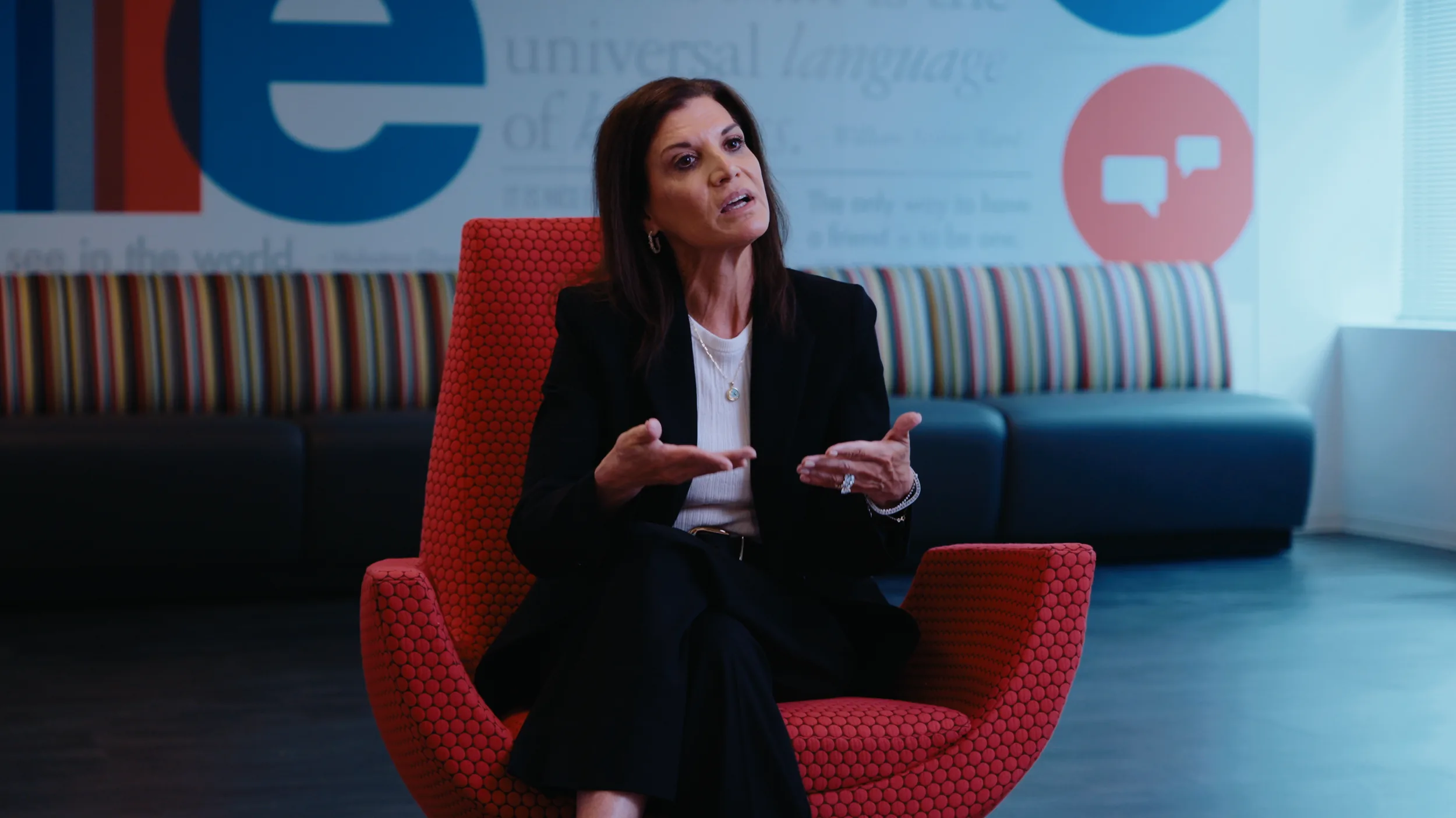 A woman with dark hair in a black suit speaks with open hands from a red chair in front of a mural wall and striped bench
