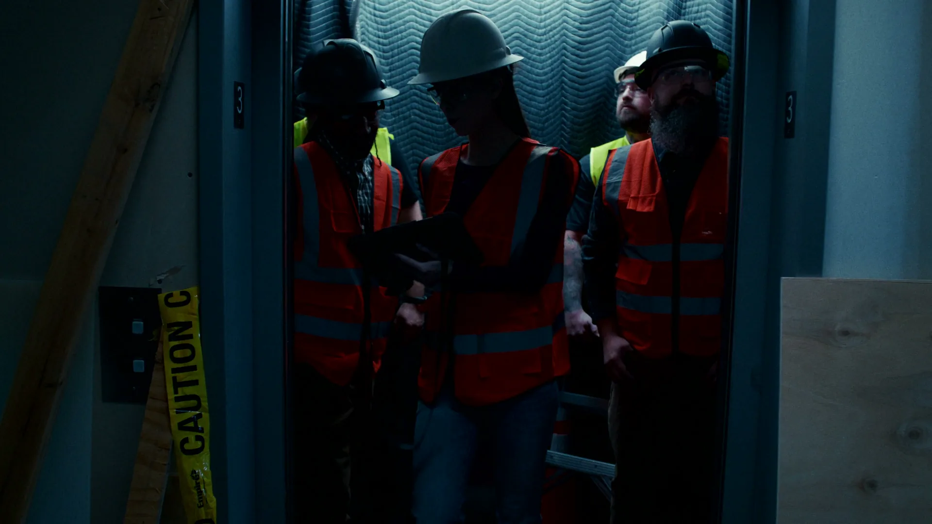 Delivered frame: a construction crew in hard hats and orange safety vests in an elevator car, one checking a tablet
