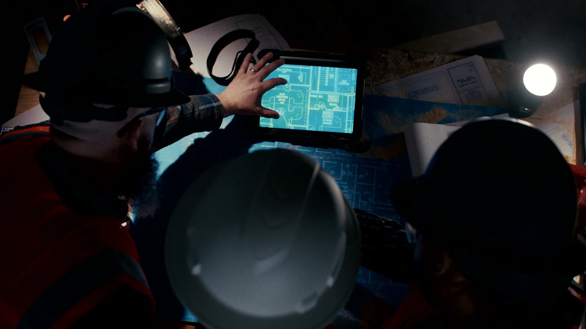 Delivered overhead frame: crew in hard hats reviewing plans on a tablet over blueprints