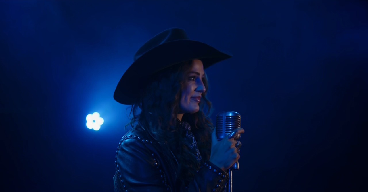 Kat Hasty in a black cowboy hat smiles in profile at a vintage microphone, wrapped in solid deep blue stage light.