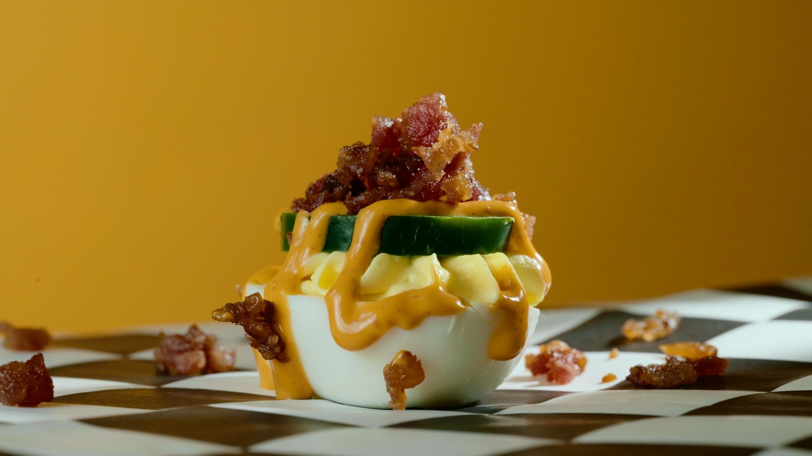 A loaded deviled egg on a black and white checkerboard against a saturated mustard backdrop.