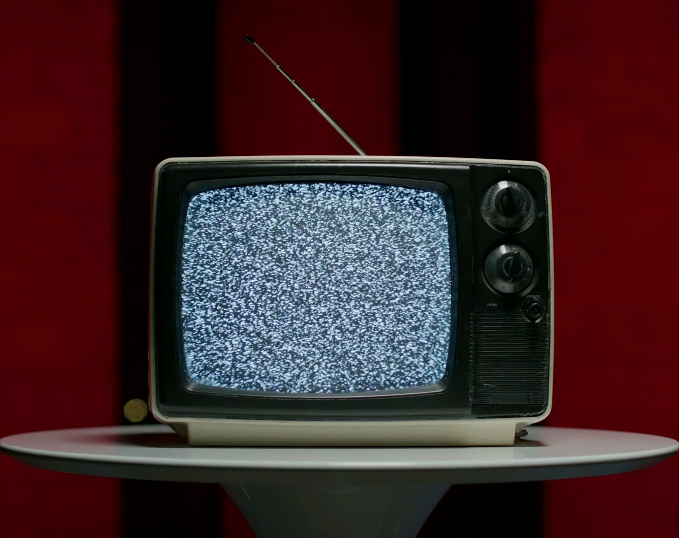 A vintage television showing static sits alone on a pedestal in front of a red curtain.
