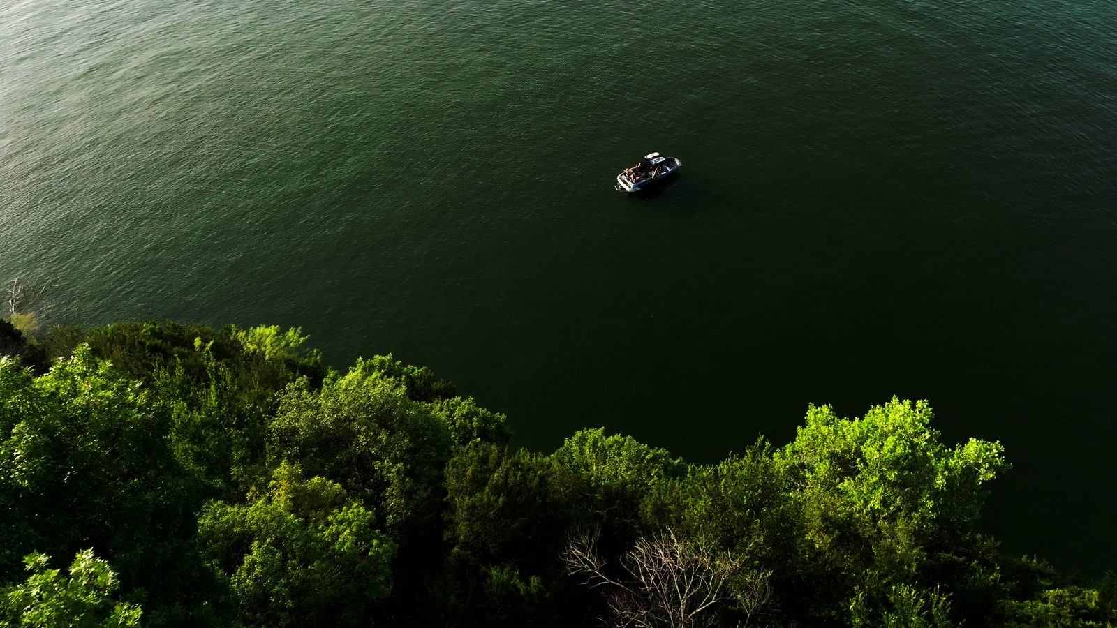 Aerial view of a single wakeboat small on a wide field of dark green lake water above a tree line.