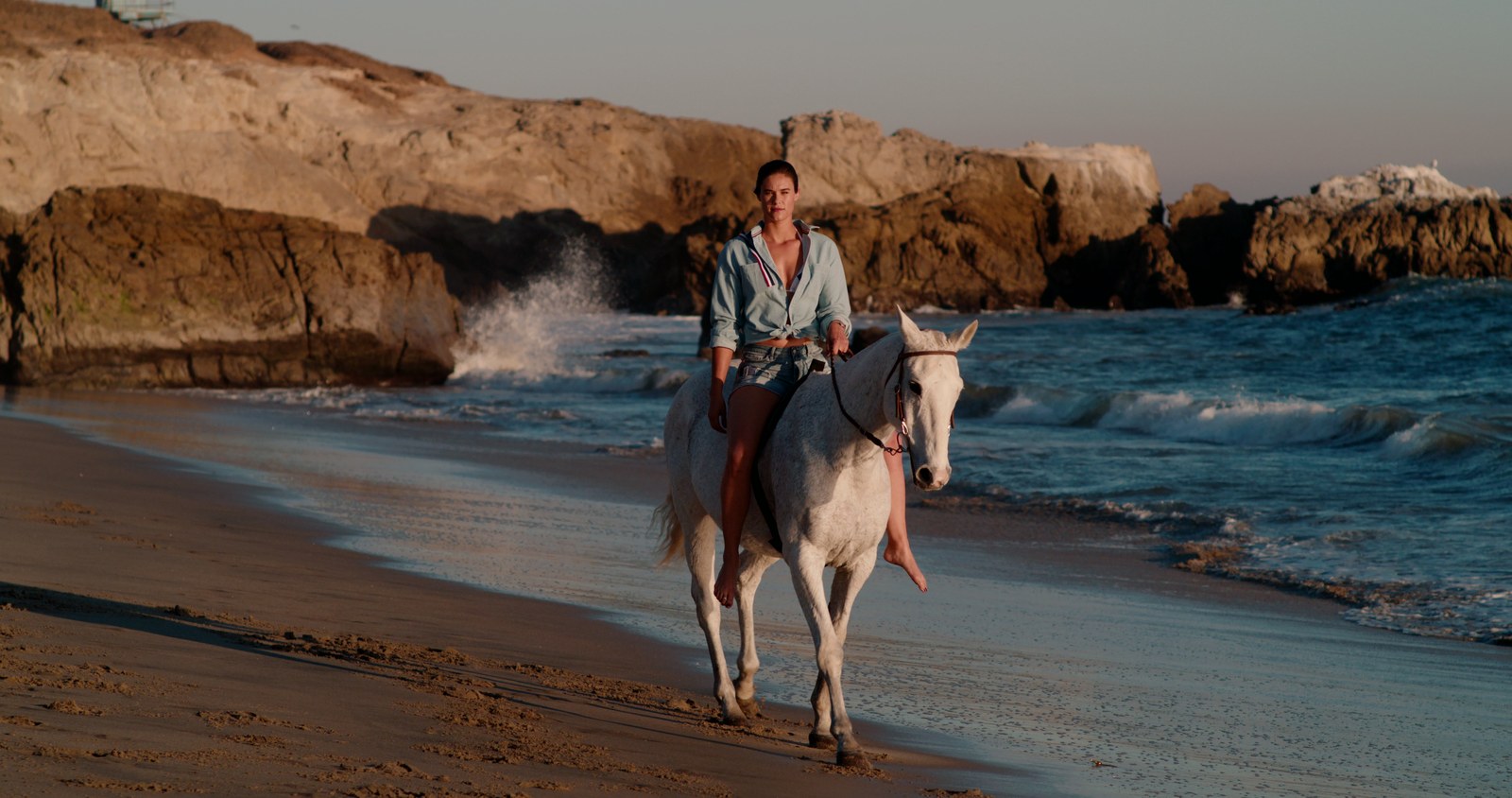 A rider on a white horse walks the surf line at golden hour as a wave breaks against the rocks behind her.