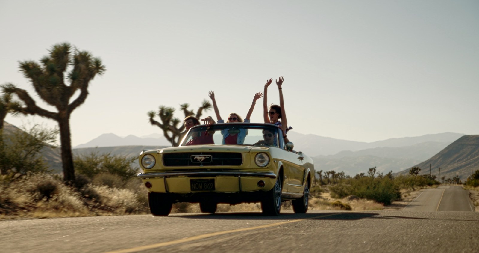 A pale yellow vintage Mustang convertible drives a desert road past Joshua trees, everyone in the back with arms raised.