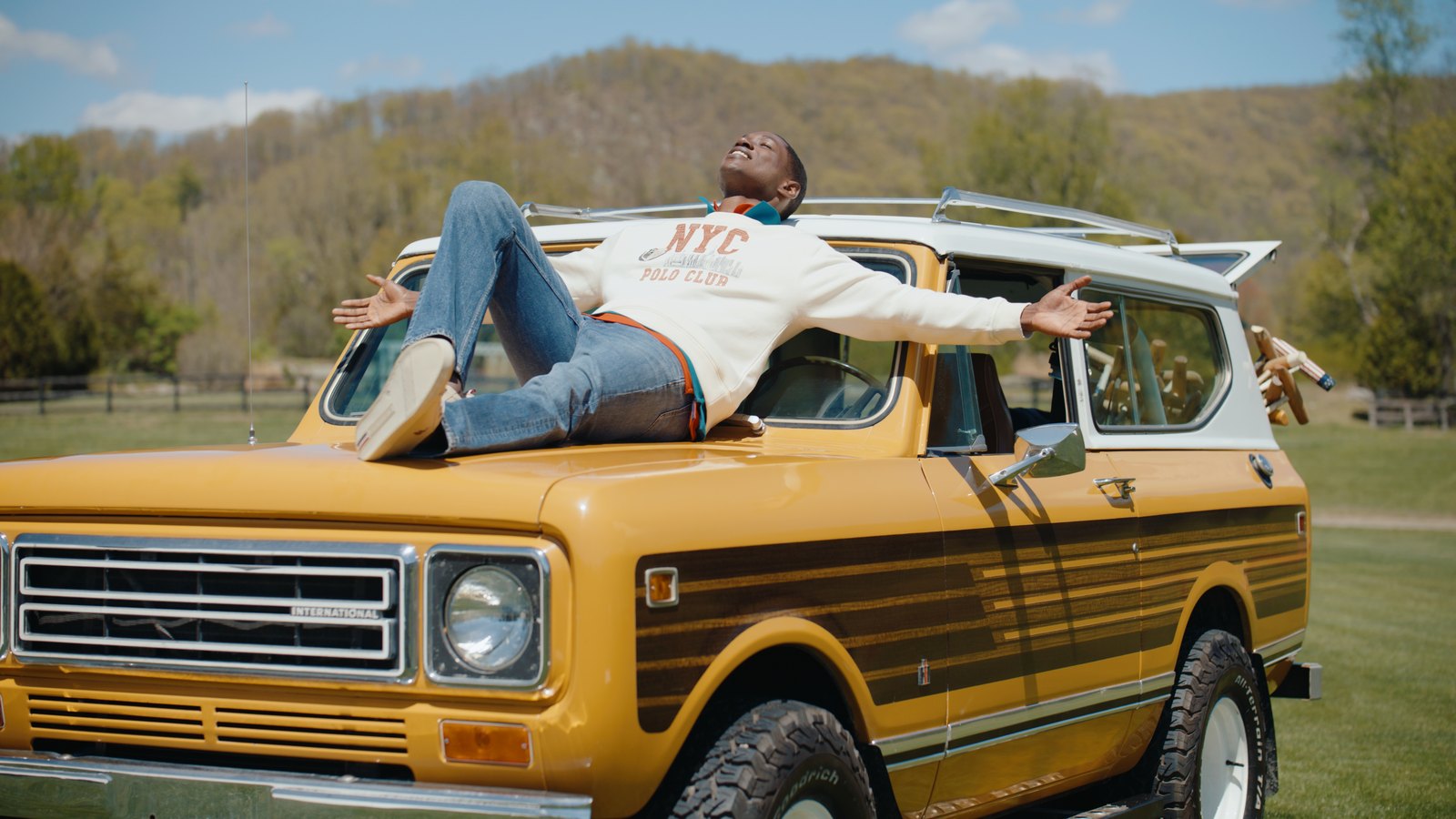 A man stretches out with open arms across the hood of a yellow vintage International Scout in a green field below wooded hills.