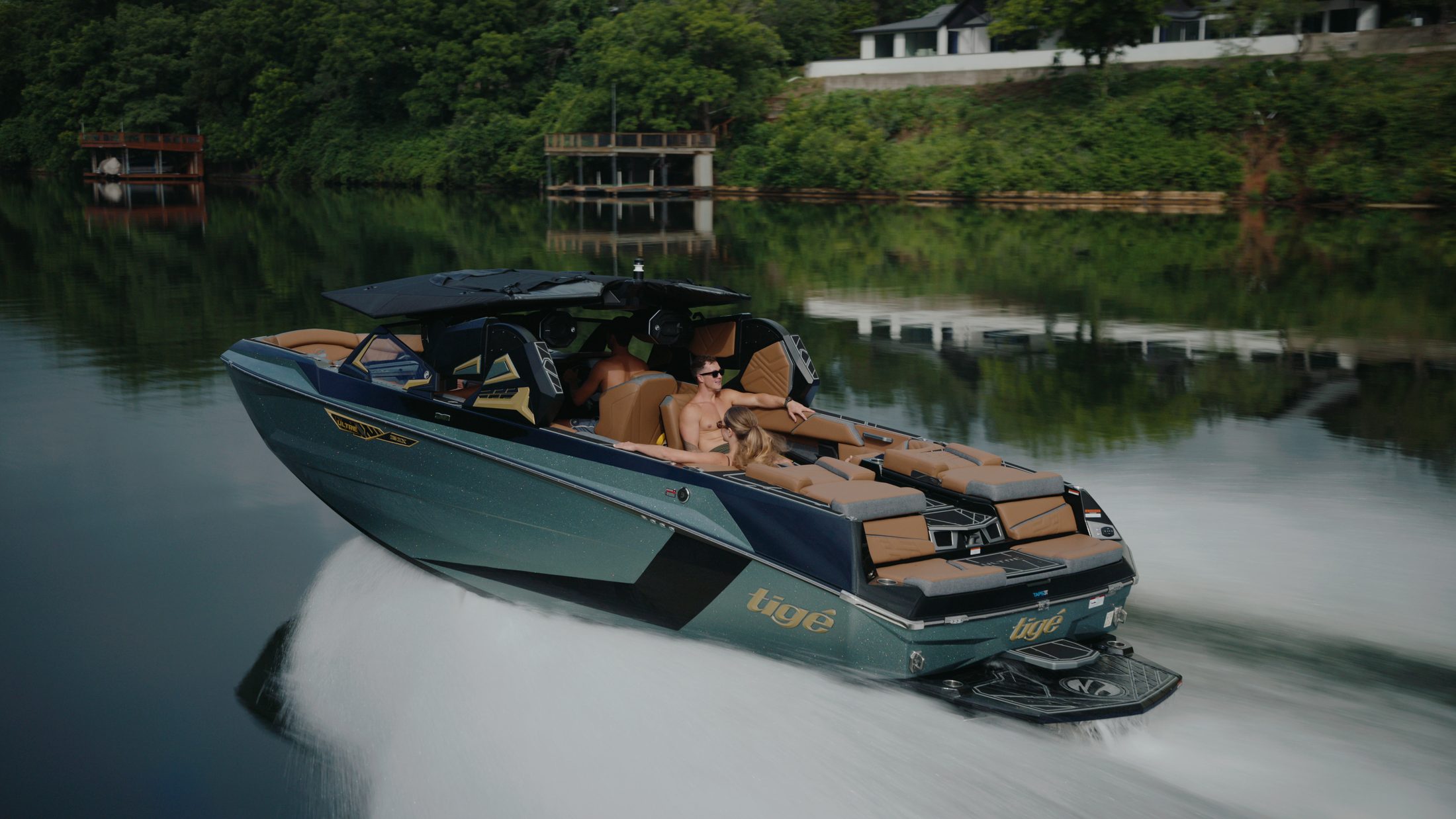 A gray and white wakeboat carves low across dark green lake water, its hull catching warm light against a wooded shore.