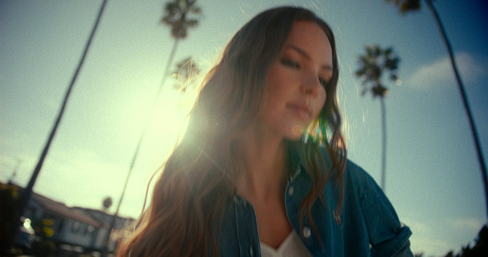 Shot from below, a woman in a denim shirt looks down as the sun burns through her hair, palm trees overhead.