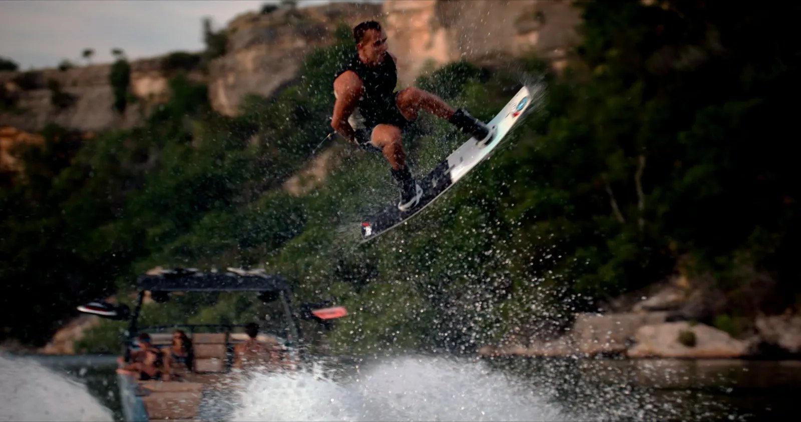 A wakeboarder flies above the wake as low sun backlights the spray against dark cliffs and trees.
