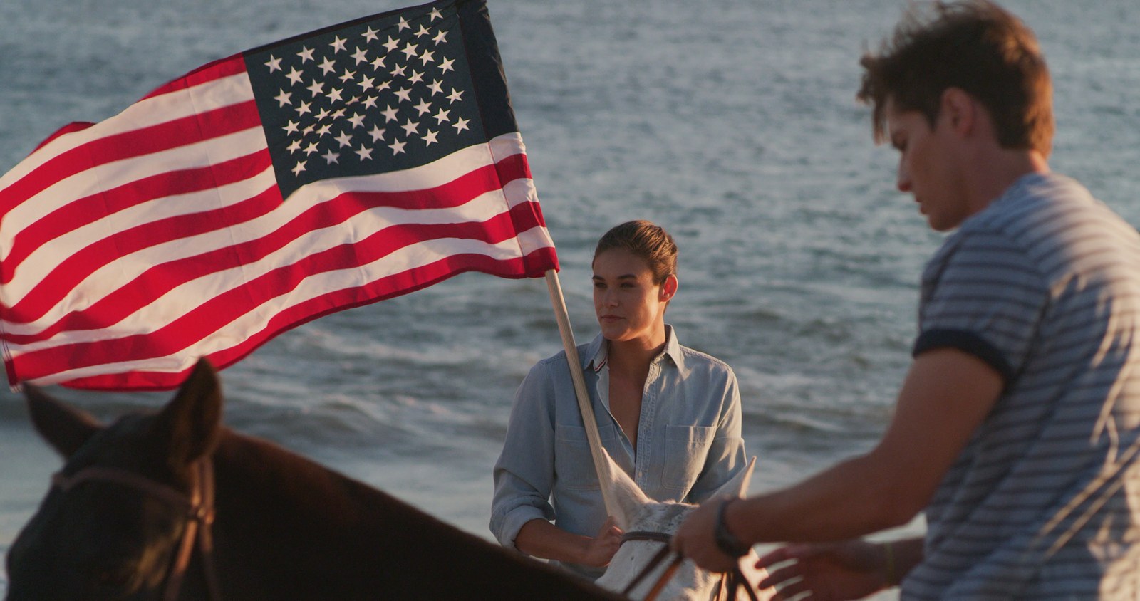 A woman on horseback holds an American flag snapping in the wind against the ocean at golden hour.