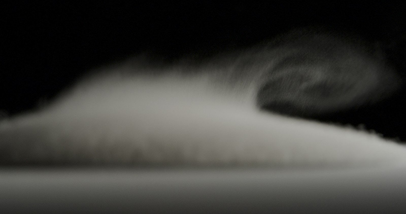 Powder lifts off a speaker and curls into the air like a breaking wave against black.