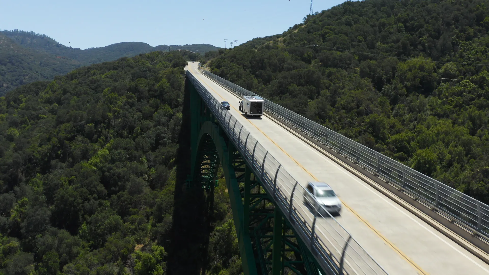 Aerial of a truck towing a Living Vehicle trailer across a green steel arch bridge