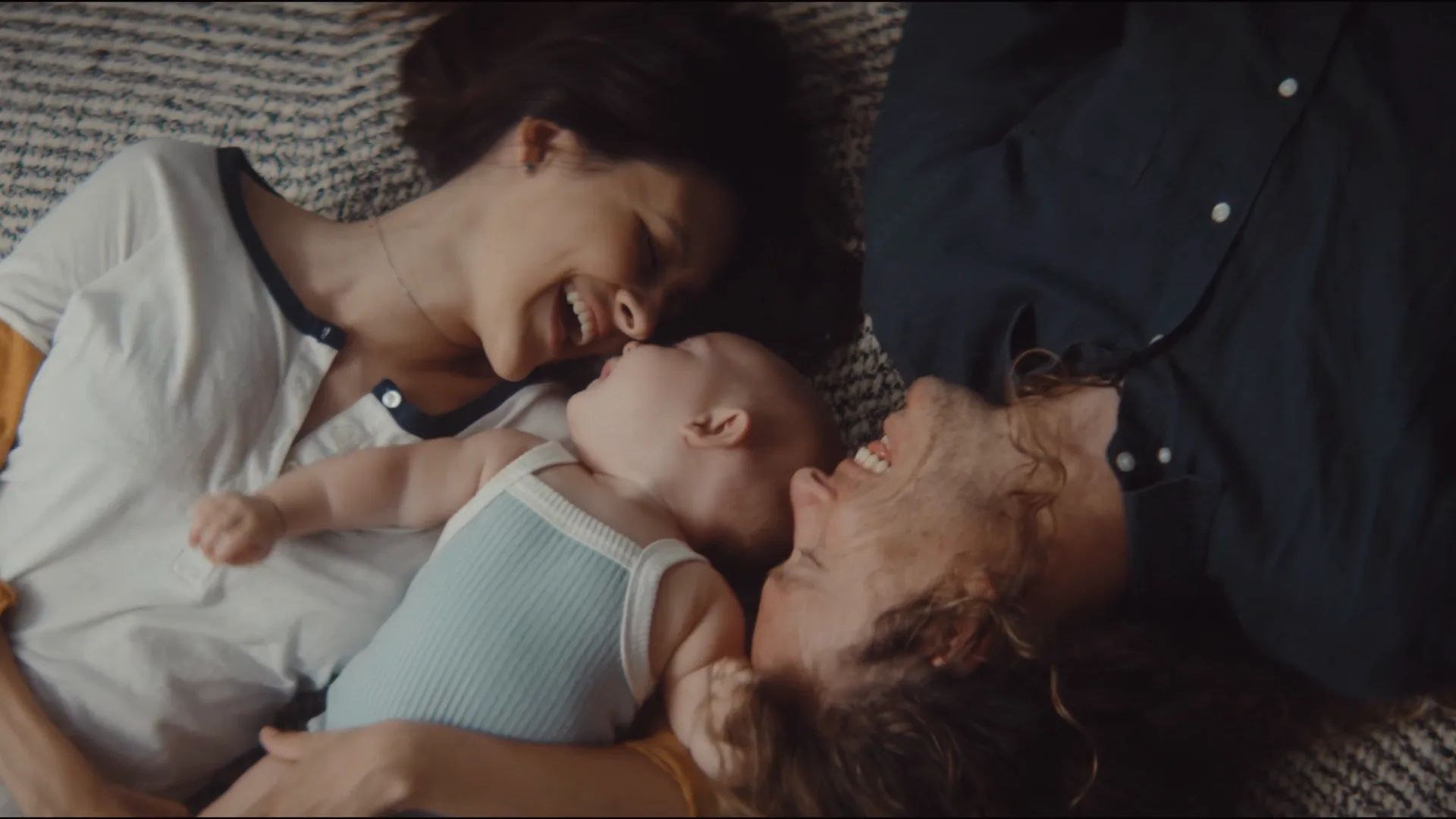 Parents lying on a rug, leaning in to kiss their laughing baby