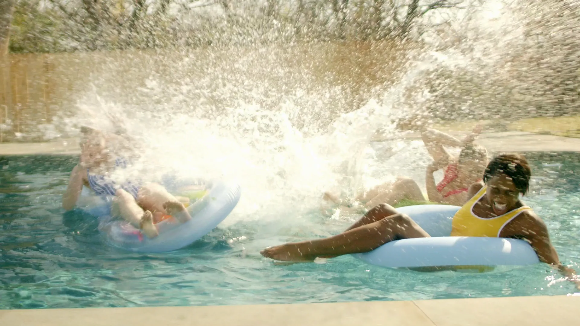 Friends splashing on inflatable floats as a wall of water bursts across a pool