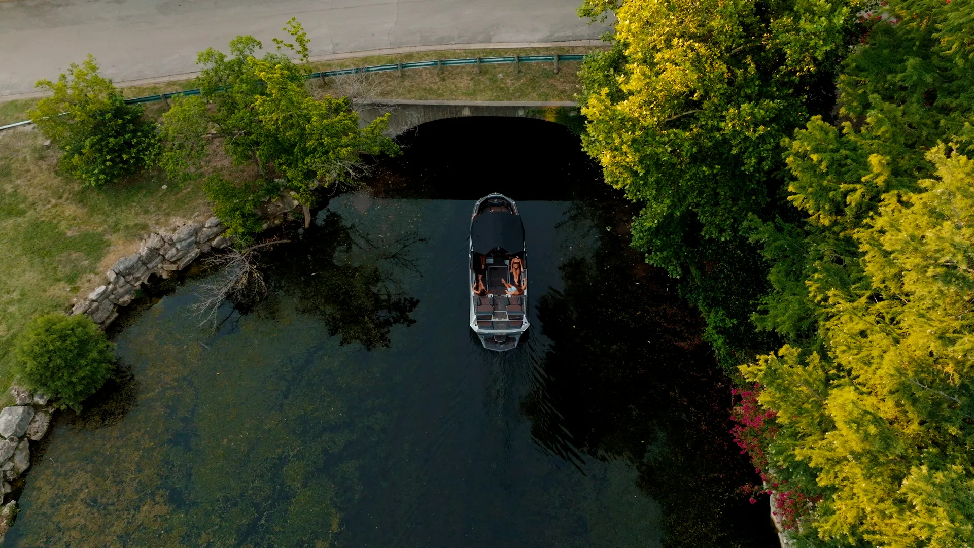 Top-down drone on a Tige boat in a narrow tree-lined channel by a stone bridge