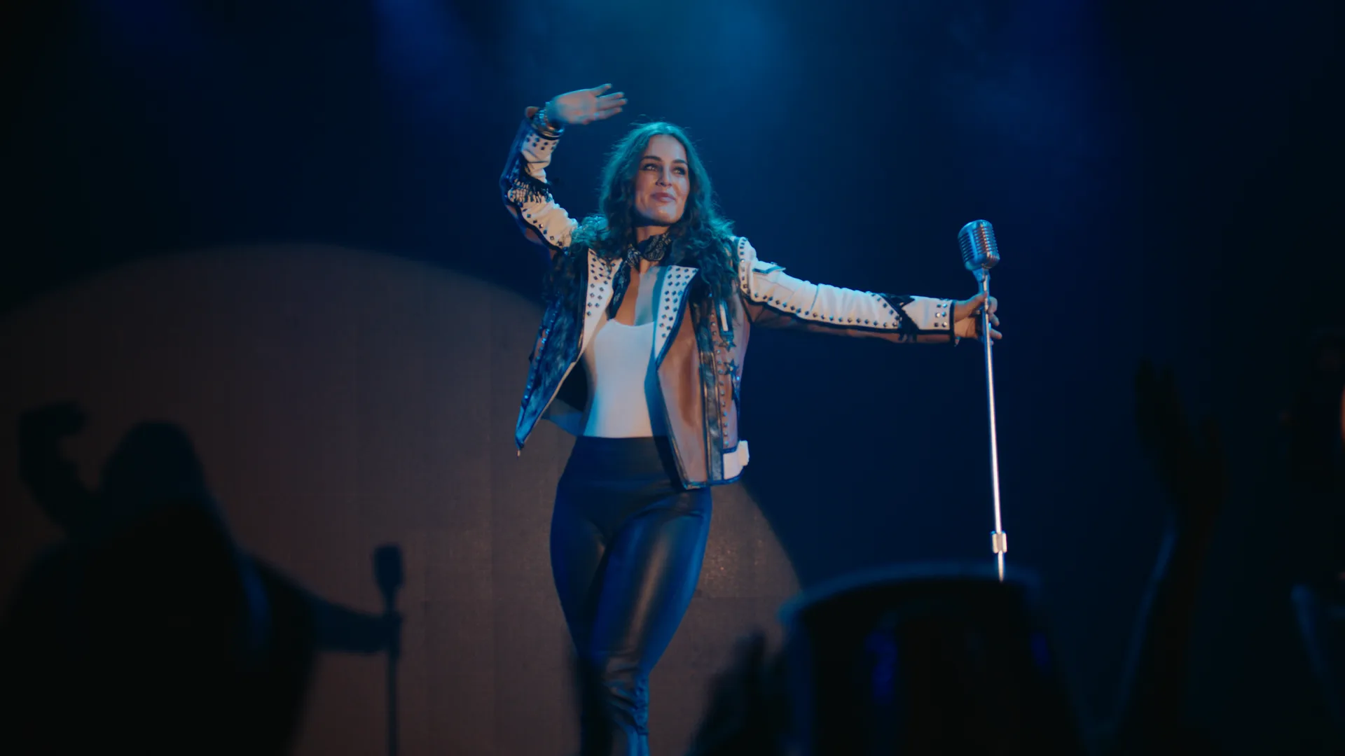 Kat Hasty waving from a dark stage, mic stand in hand