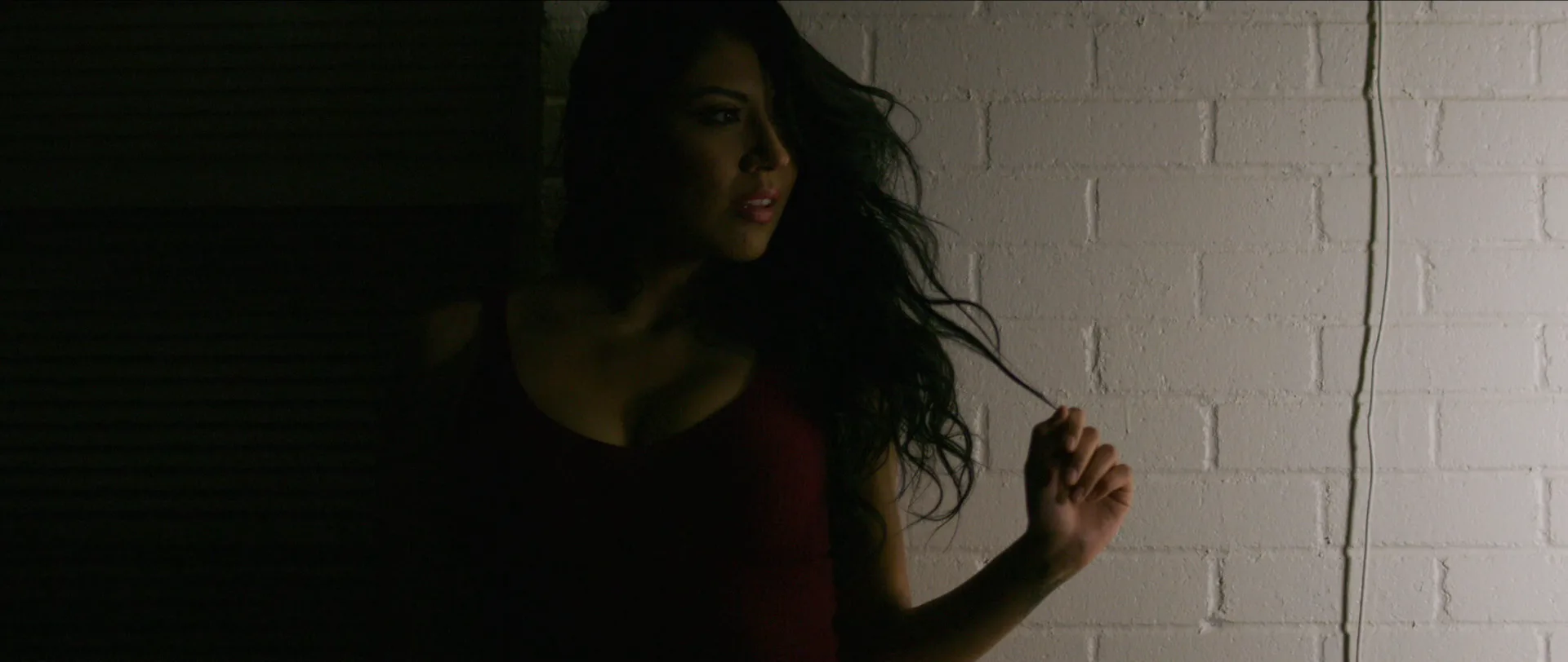 A woman against a white brick wall in low light, hand in her hair