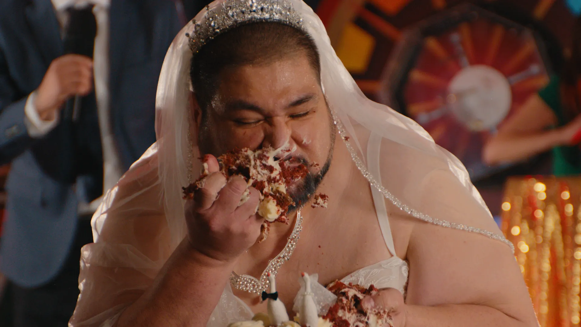 A man in a wedding dress and veil smearing red velvet cake on his face