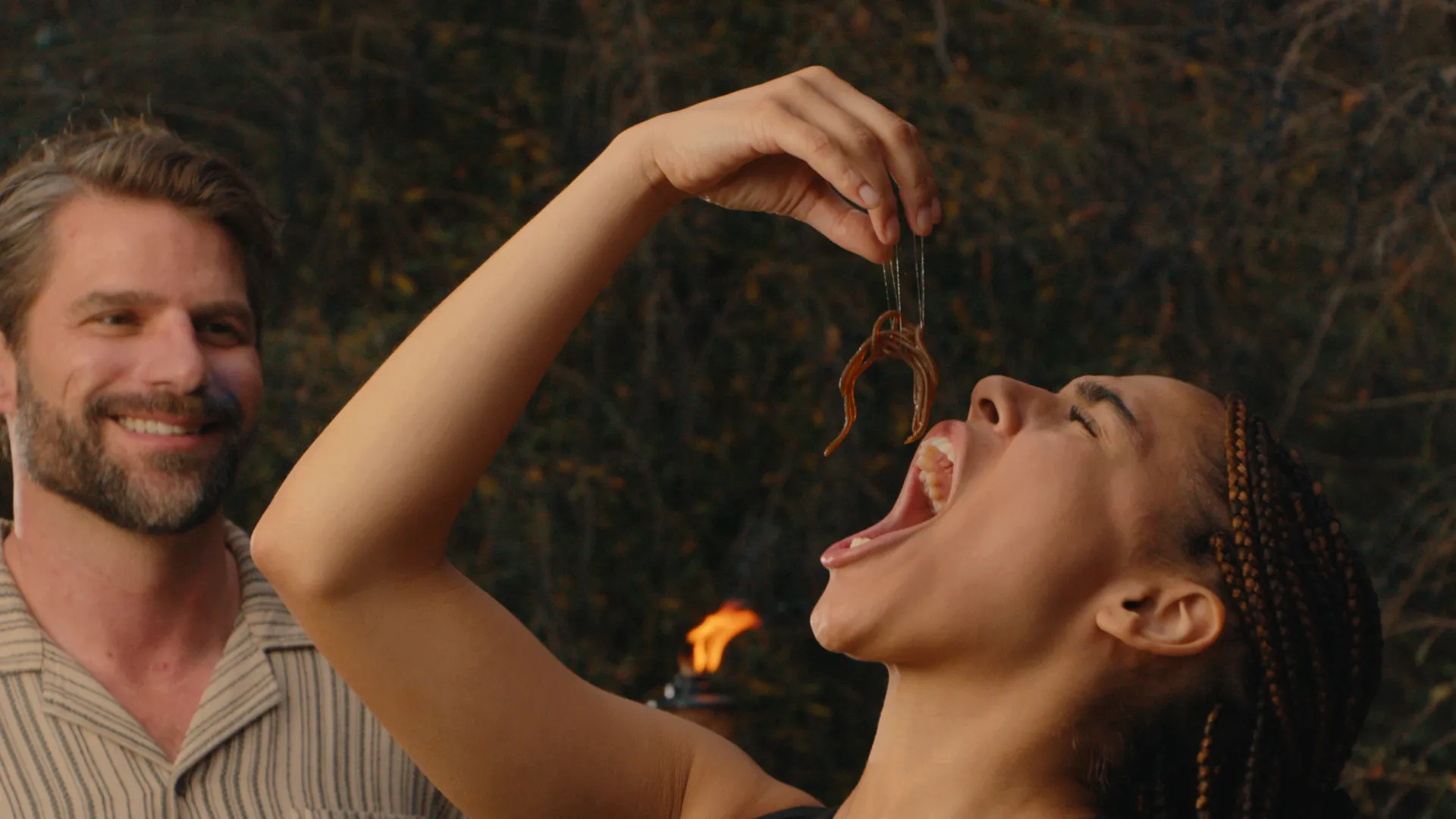 A woman dropping a worm into her mouth as a man grins beside her