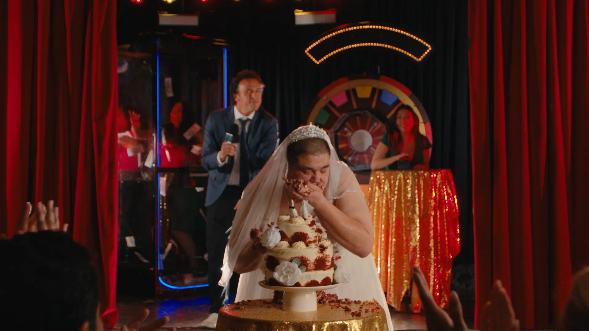 The man in the wedding dress on a red-curtained game-show stage under arcade lights