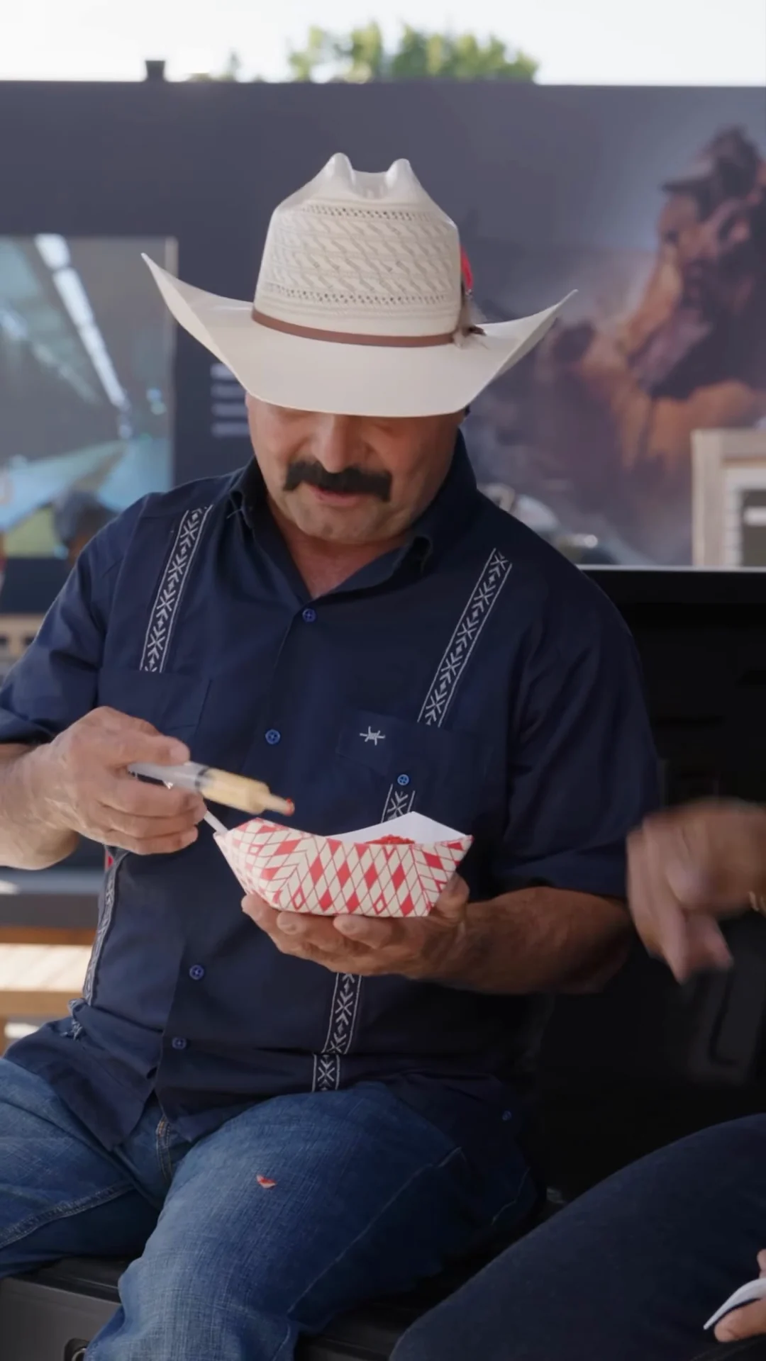 A reviewer in a straw hat taking a bite of fair food on the tailgate