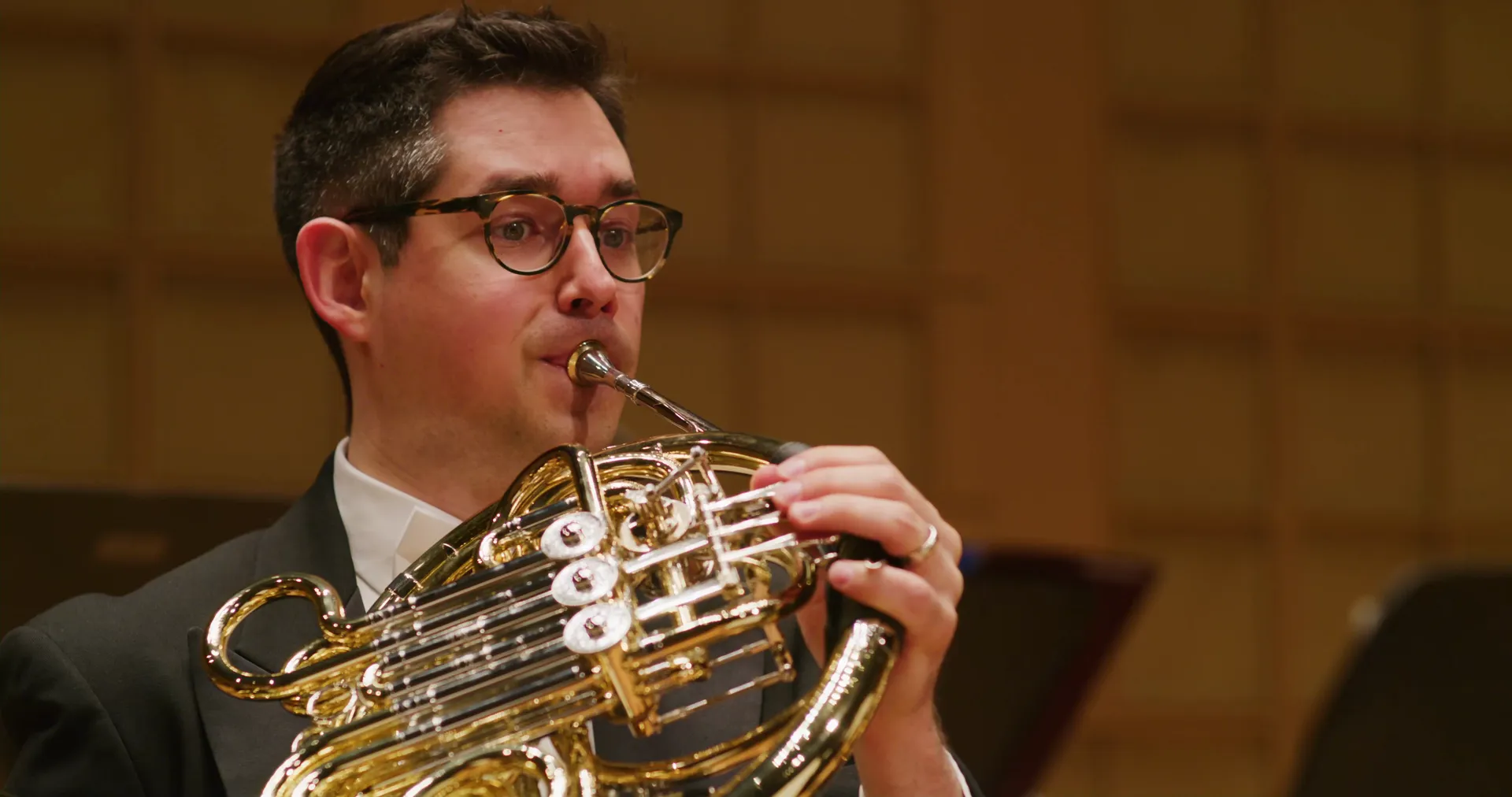 A French horn player in glasses mid-phrase
