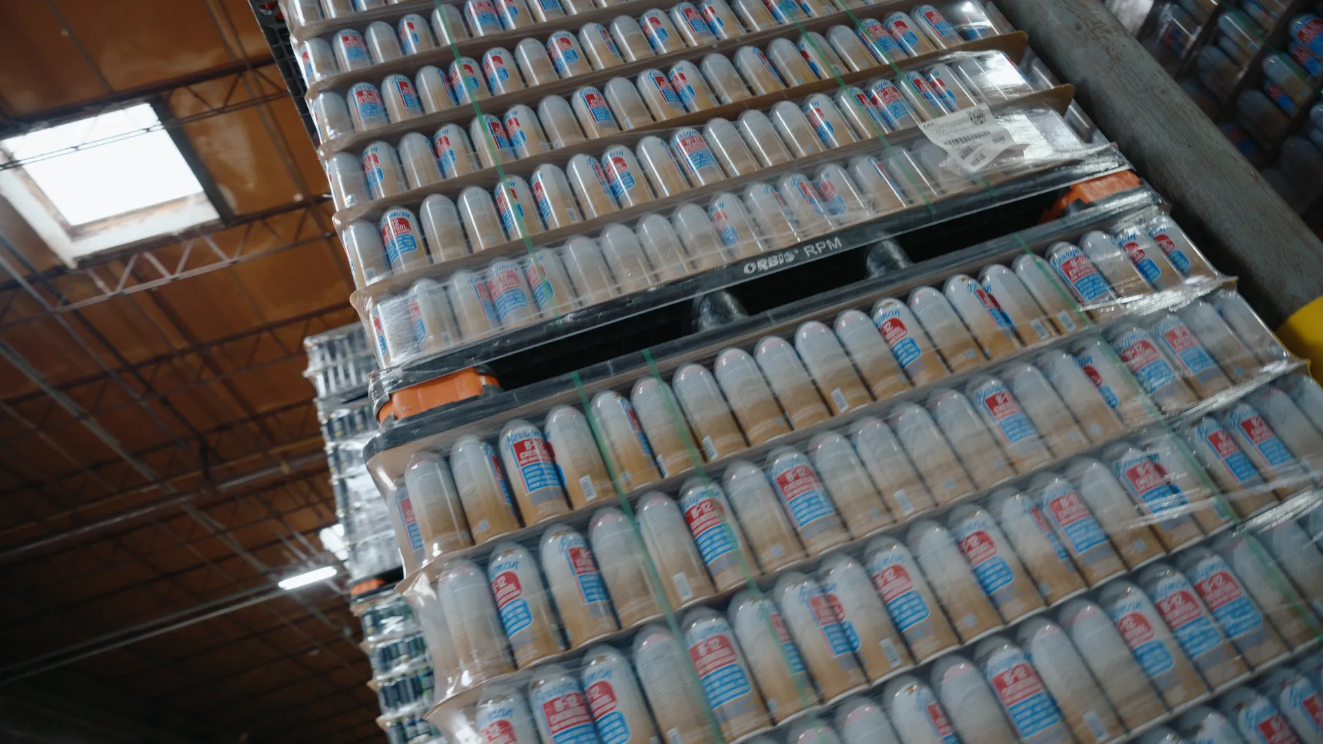 Low angle up a tower of shrink-wrapped pallets of aerosol cans under a warehouse skylight