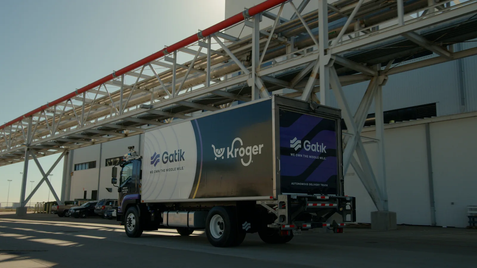 Camera car tracking a Gatik box truck under a steel truss walkway beside an airport hangar