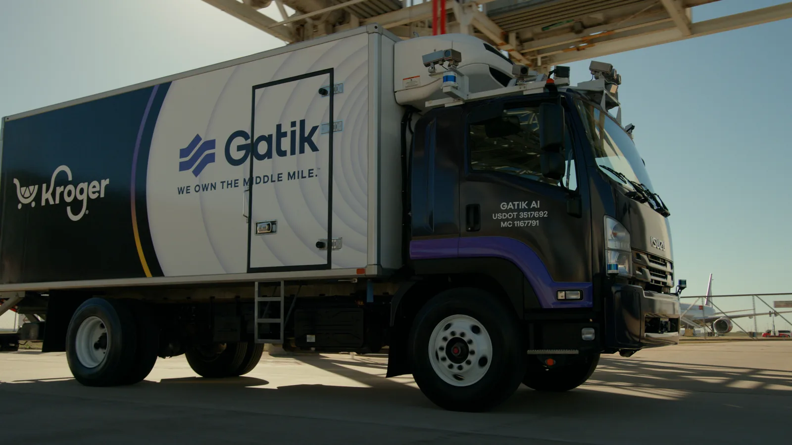 Low three-quarter tracking shot of a Gatik x Kroger truck under a hangar truss
