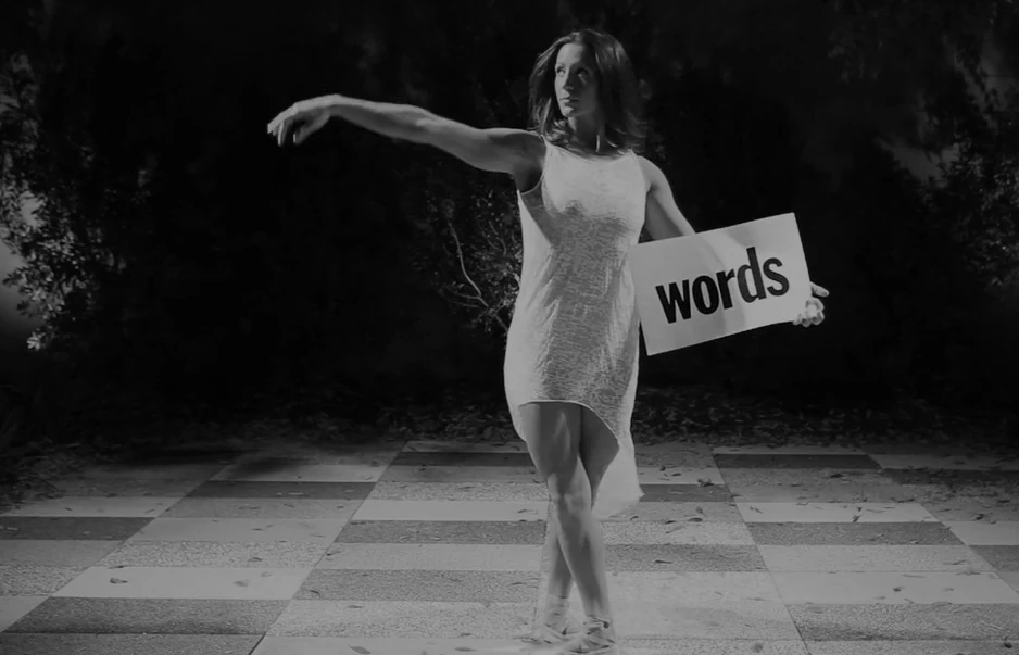 A dancer in a white dress reaches out across a checkered courtyard at night, holding a sign reading words