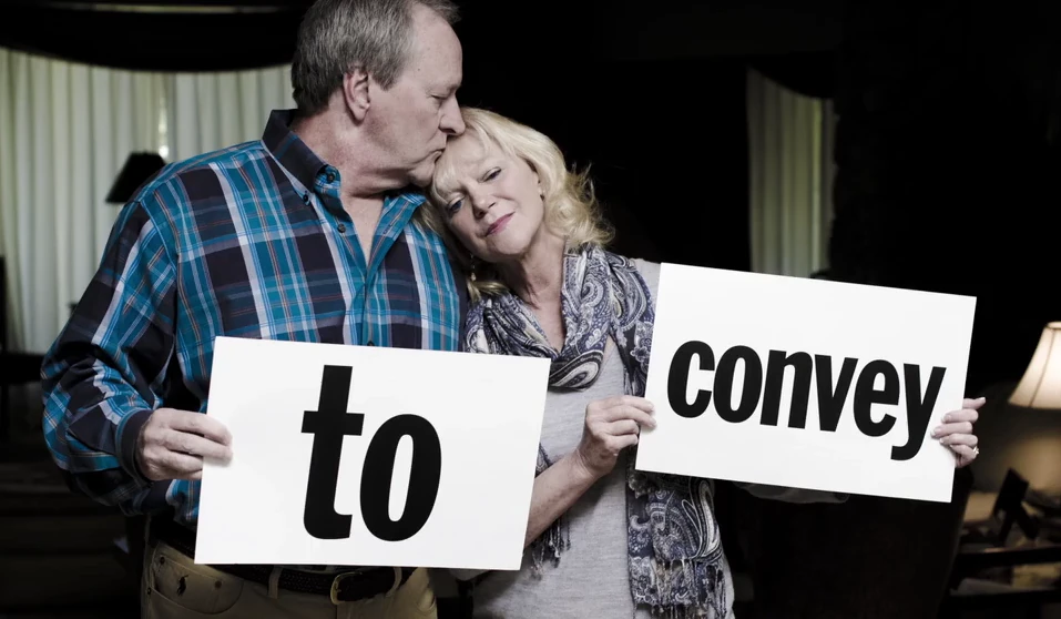 A husband kisses his wife's head as they hold signs reading to and convey