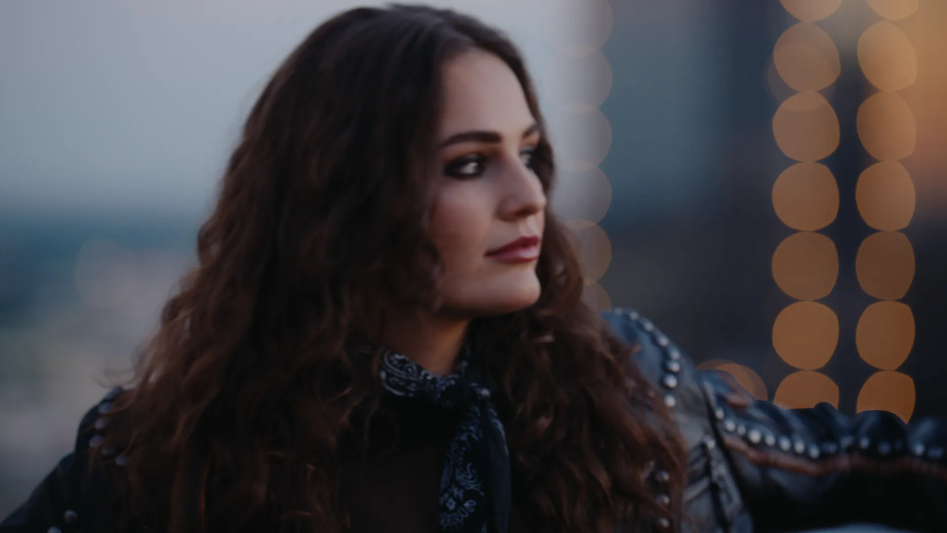 Khasty in a studded jacket on a rooftop, city lights soft behind her