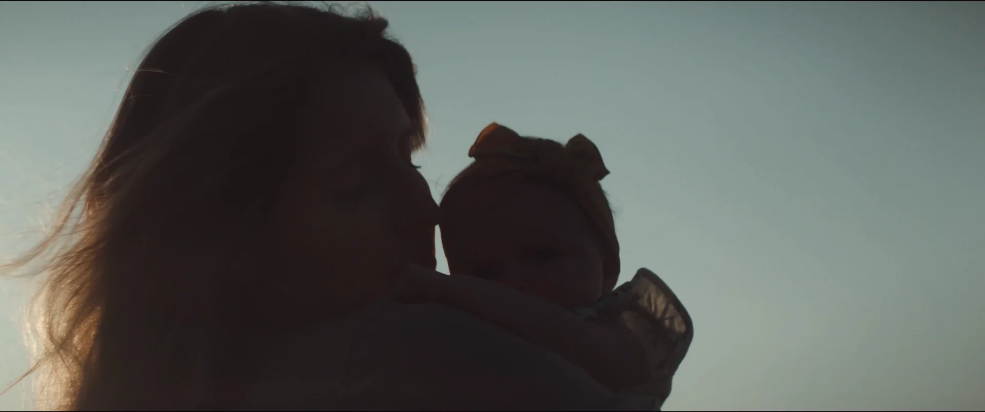 A mother kissing her baby daughter’s head, both silhouetted against a pale sky on a lake dock