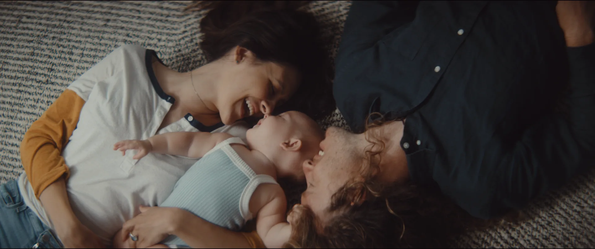 Parents lying on a rug, both leaning in to kiss their laughing baby