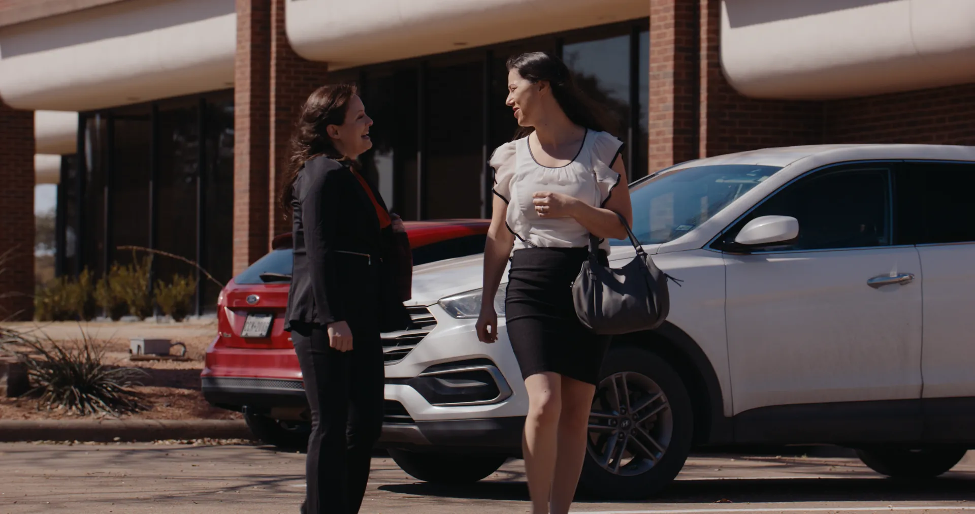 Two women talking beside parked cars outside an office