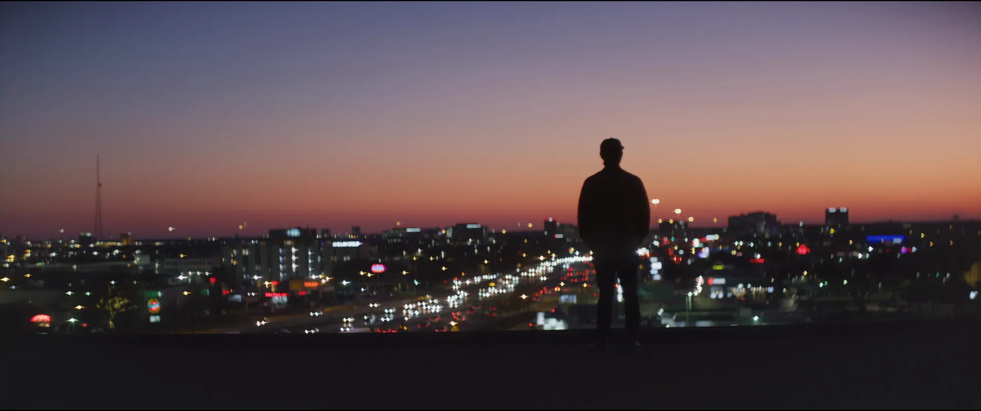 A lone figure silhouetted on a rooftop against an orange and violet dusk sky, city lights spread out below