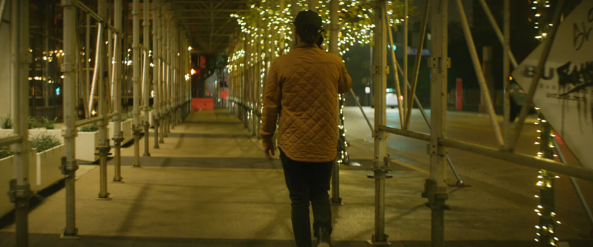 A man walking a covered downtown sidewalk under scaffolding wrapped in string lights