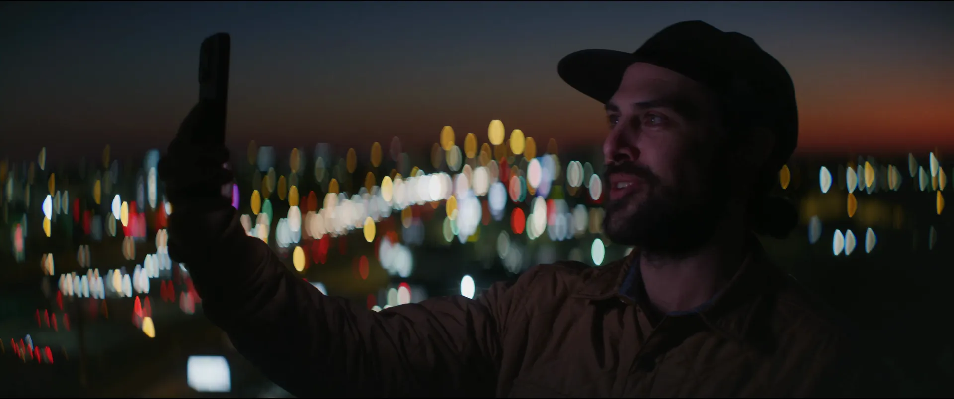 A smiling man holding his phone up for a photo, the city behind him melted into bokeh