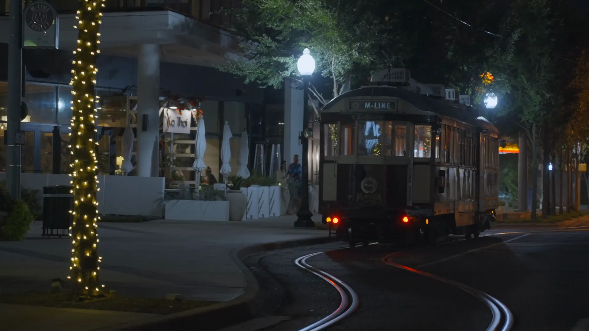 An M-Line trolley rounding a curve at night past a tree wrapped in string lights