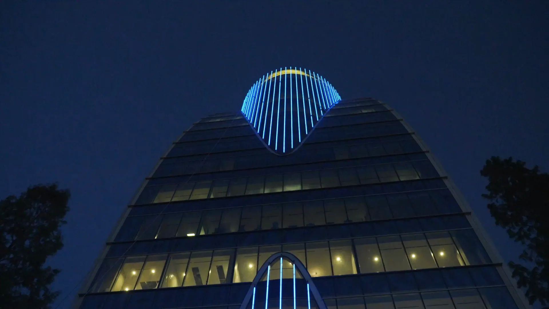 A glass office tower crowned with a blue-lit arch at blue hour