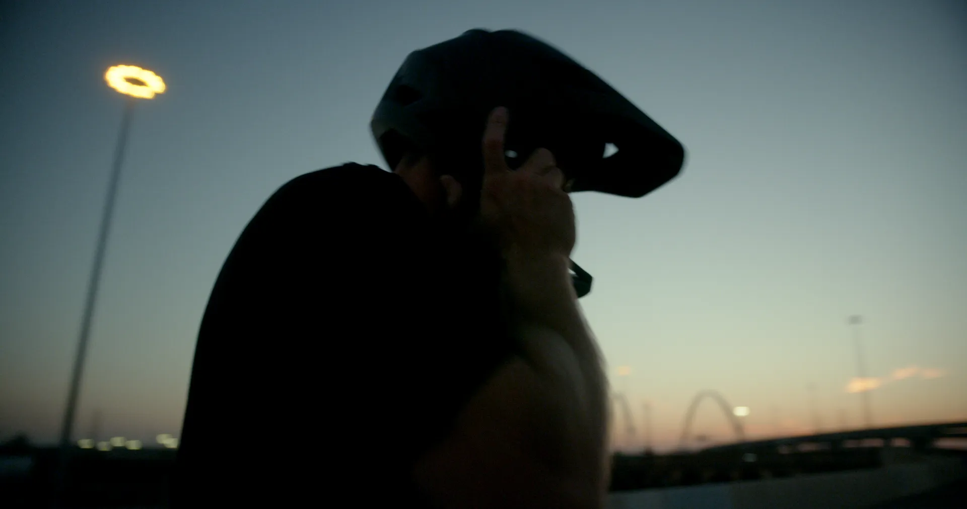 A rider’s helmeted silhouette against a dusk sky, the Margaret Hunt Hill Bridge arches in the distance