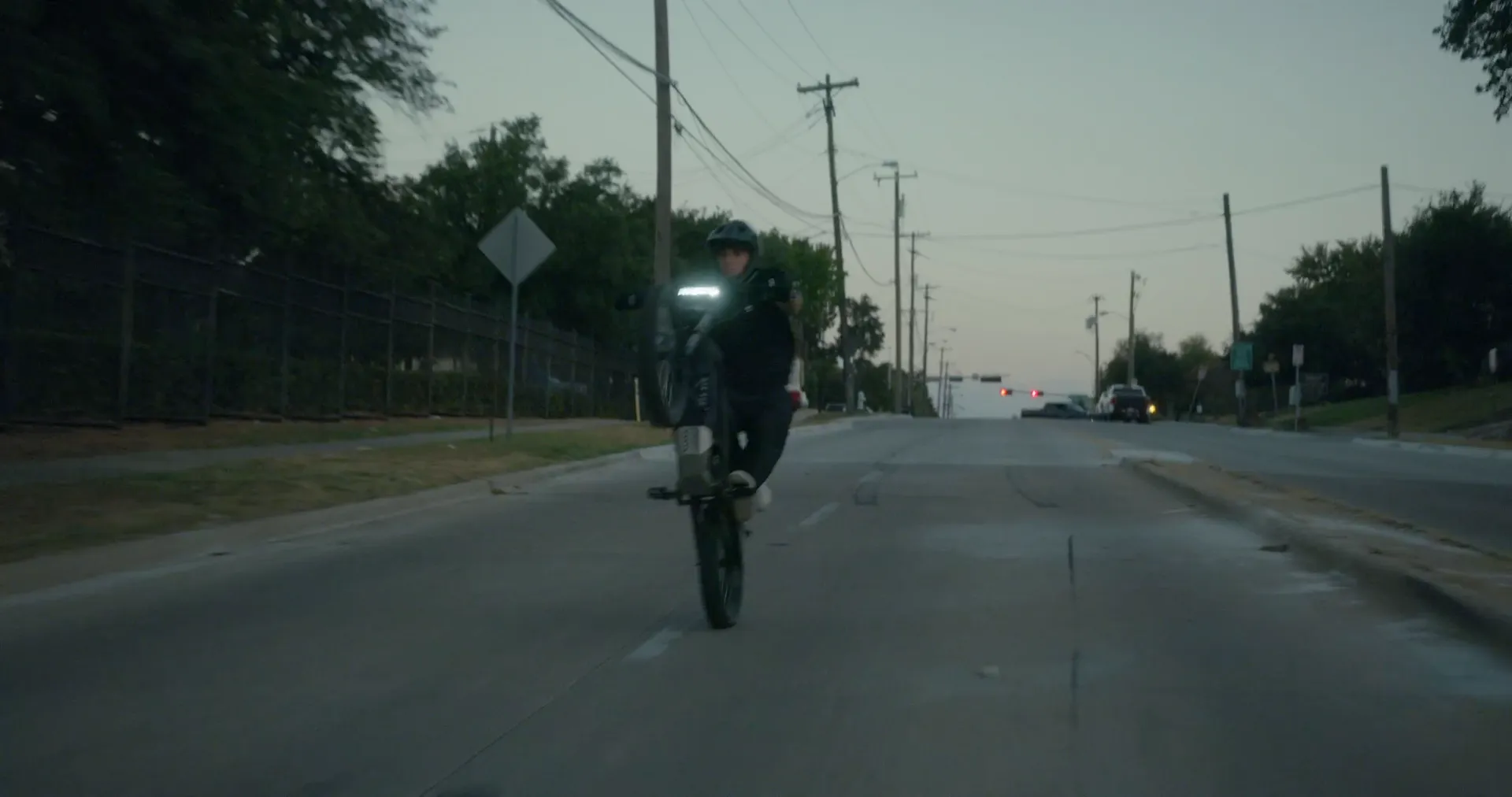 A rider wheelieing down a tree-lined street at dusk