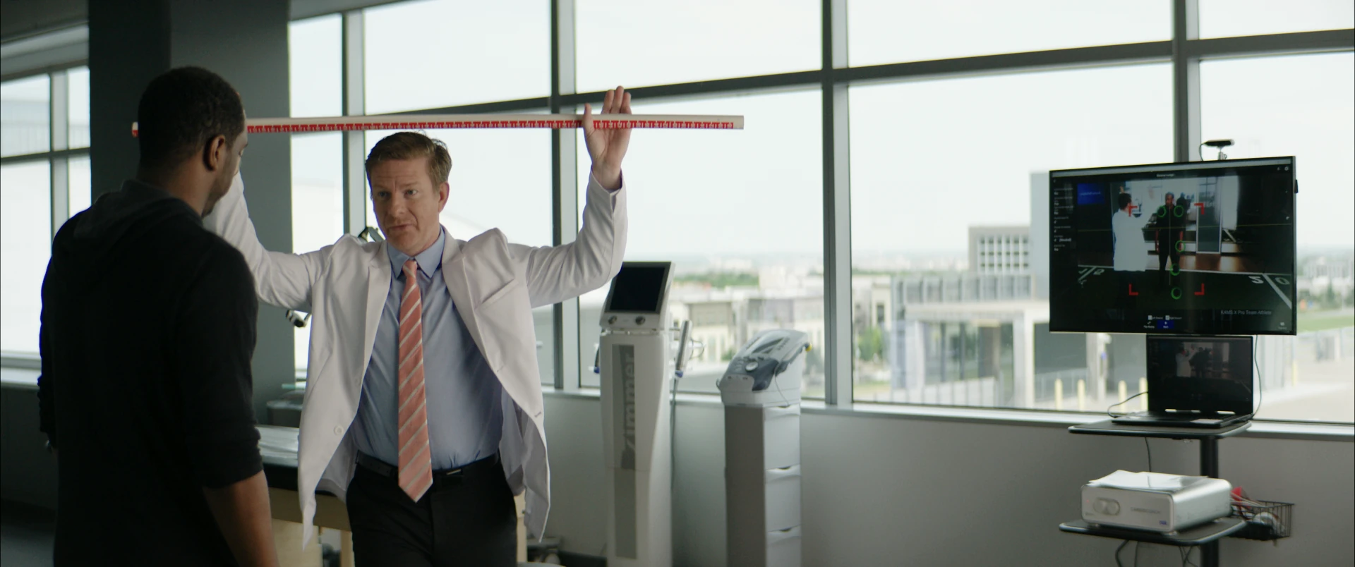 Wide shot of a doctor demonstrating an overhead dowel stretch in a sunlit clinic