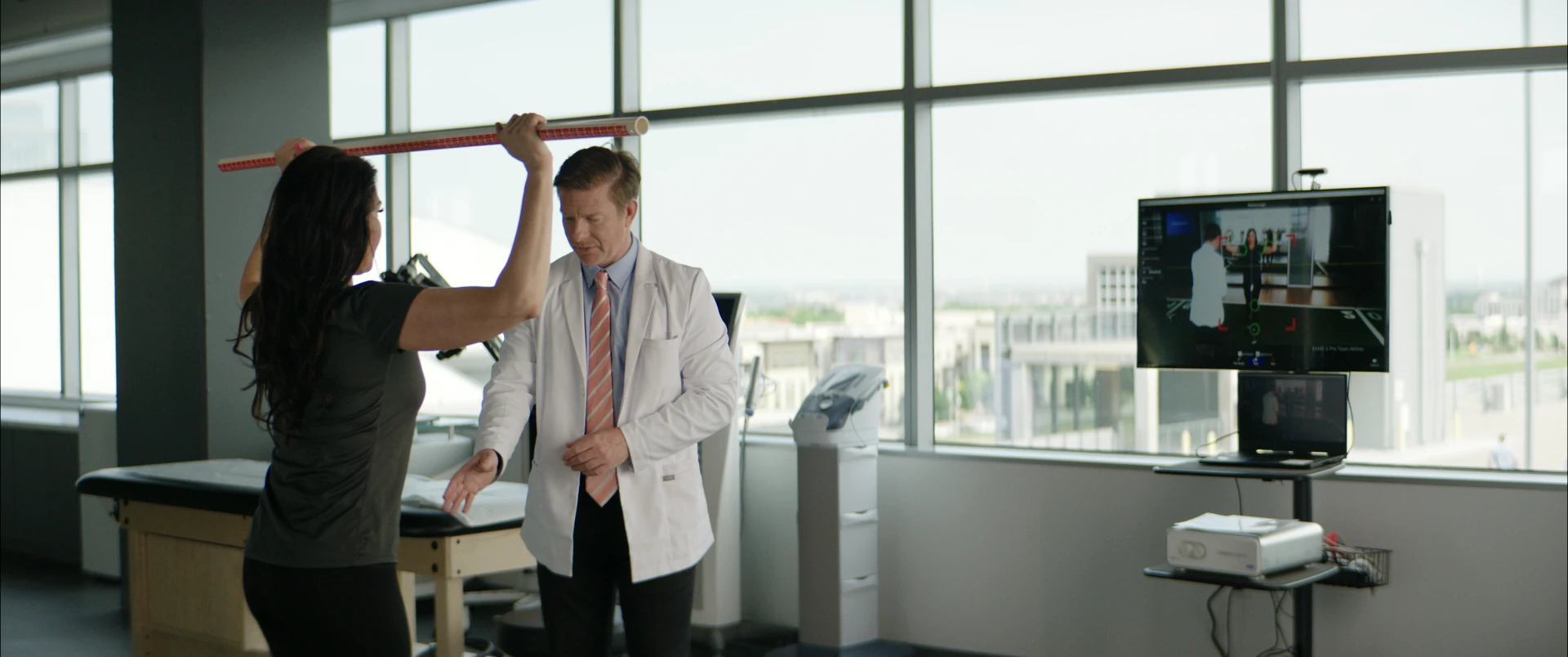 Wide shot of a patient stretching with a dowel as her doctor looks on by the windows