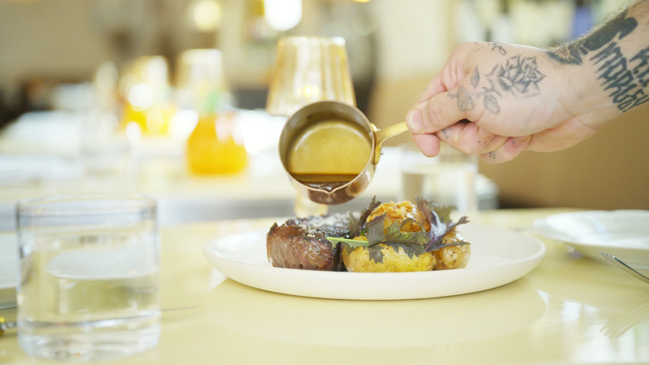 Tattooed hand pouring jus from a copper pot onto a plated steak in the dining room