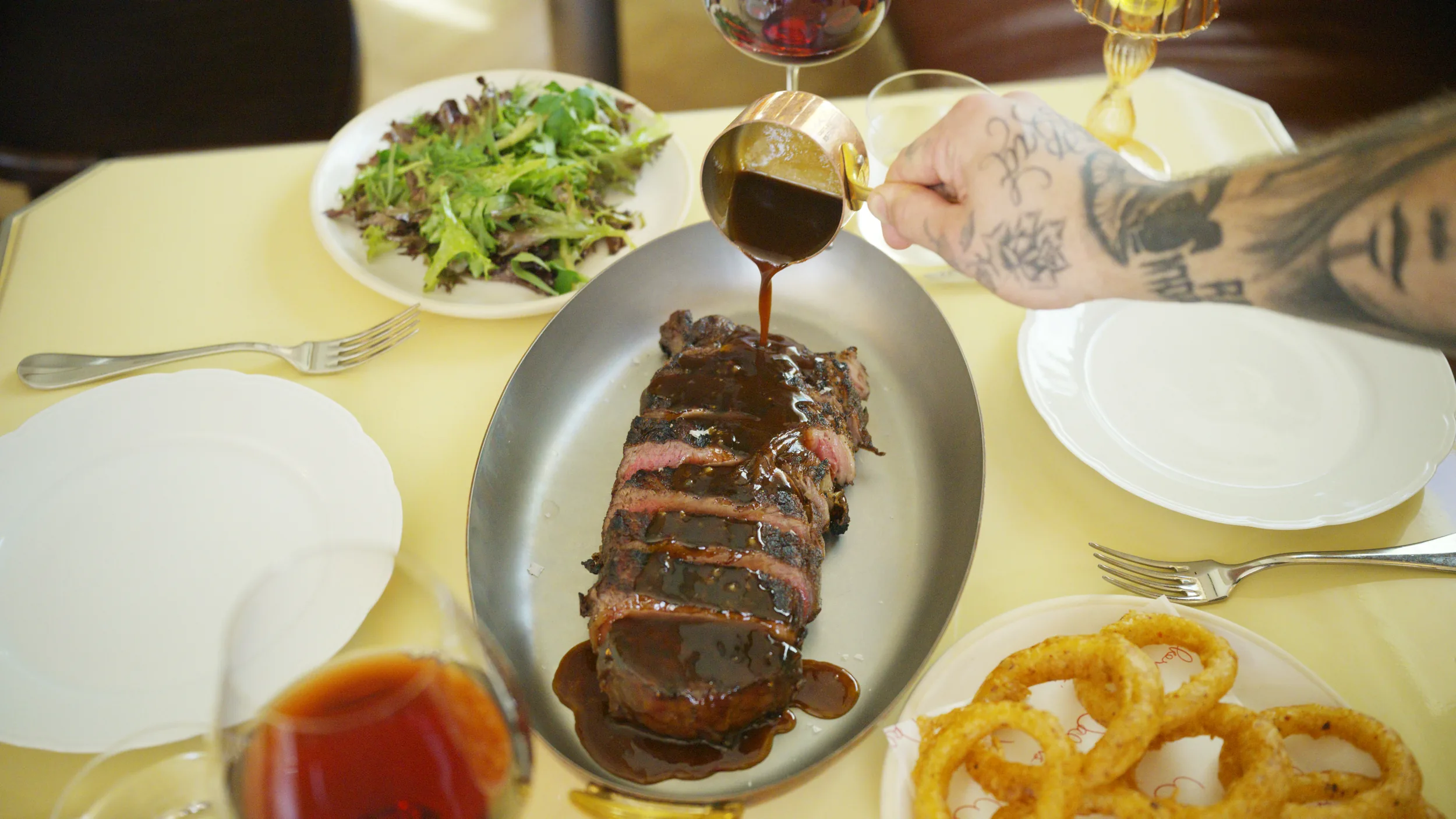 Overhead of a steakhouse table as jus pours over sliced wagyu, salad, and onion rings