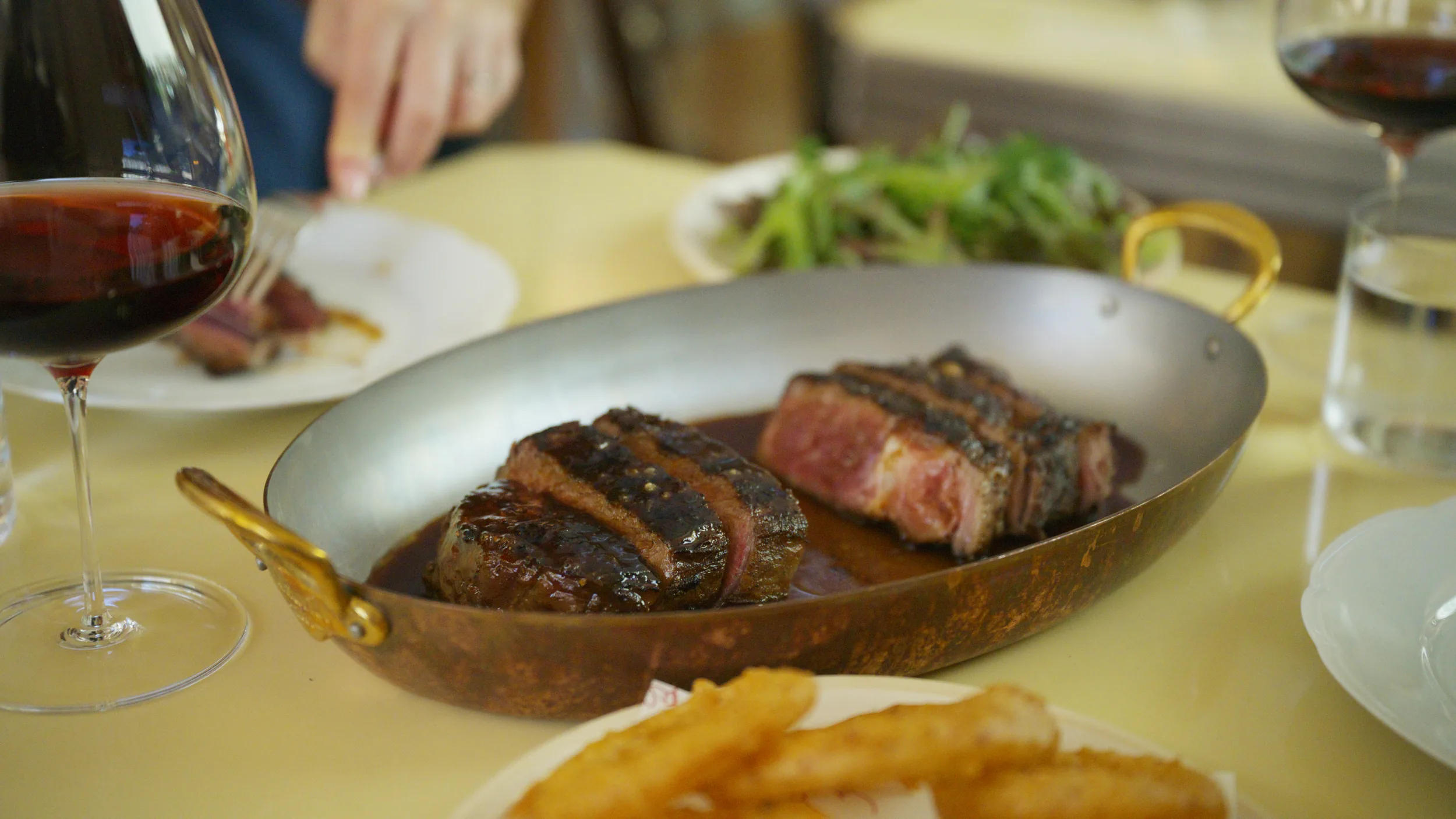 Sliced wagyu in a copper oval pan beside red wine, low across the table